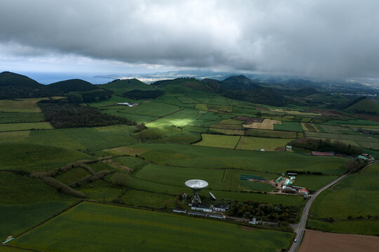 Aerial view of the radio telescope dish situated amidst the rolling green hills and agricultural fields of Sao Miguel Island, Azores, Portugal.