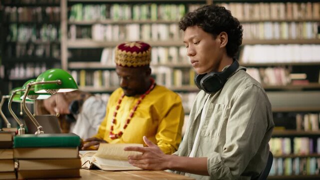 Asian student with headphones reading at library table behind books and green lamps, with another learner studying in blurred background, peers learning in study session, for online learning.