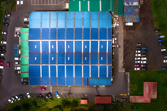 Aerial view of the Tecnovia Acores industrial facility with a large blue roof, parking areas with vehicles, and adjacent green space in Rabo de Peixe, Acores, Portugal.