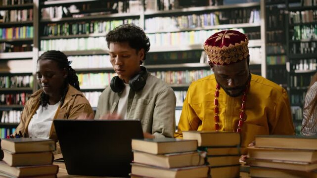 Students reviewing academic work on laptop with mature colleague at library table, surrounded by open books, stacked coursework textbooks and materials, group study, for technology research.