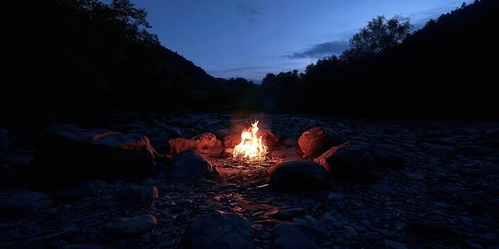 Crackling campfire illuminates a rocky riverbed under a twilight sky
