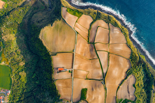 Aerial view of tilled agricultural fields with a red-roofed building and lush green vegetation bordering the Atlantic Ocean coastline in Salga, Azores, Portugal.