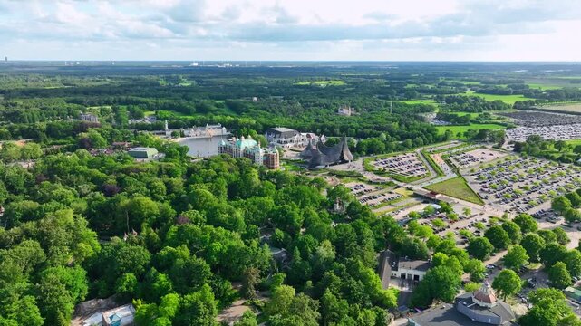 Aerial drone approaching skyline of Efteling Themepark with main entrance of in the distance surrounded by parking lot on a sunny day