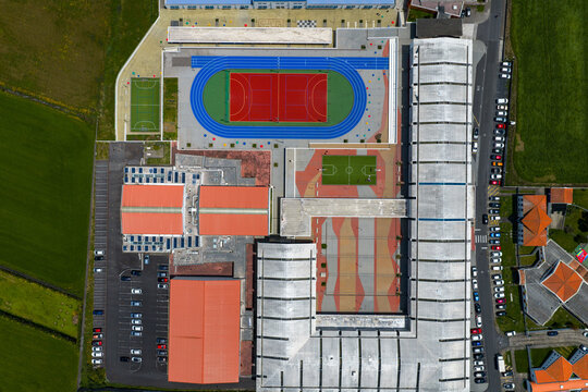 Aerial view of a school facility with a colorful sports track, soccer field, and modern buildings with orange roofs surrounded by green fields in Rabo de Peixe, Azores, Portugal.