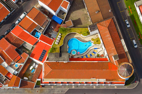 Aerial view of the Povoacao hotel featuring a uniquely shaped swimming pool, surrounded by terracotta-tiled rooftops, green spaces, and urban streets in Povoacao, Azores, Portugal.