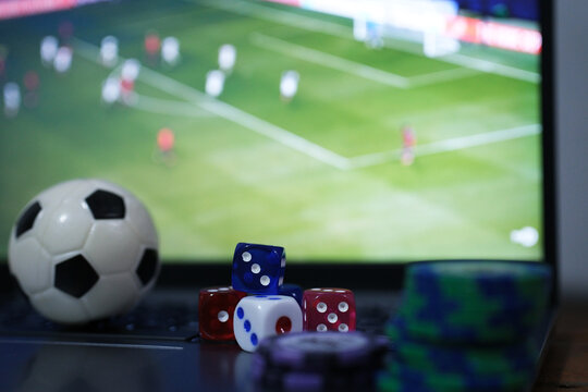 Online soccer gambling. Image of a ball, gambling tokens, and dice in front of a laptop with a live soccer broadcast. Live scores. Gambling bets.
