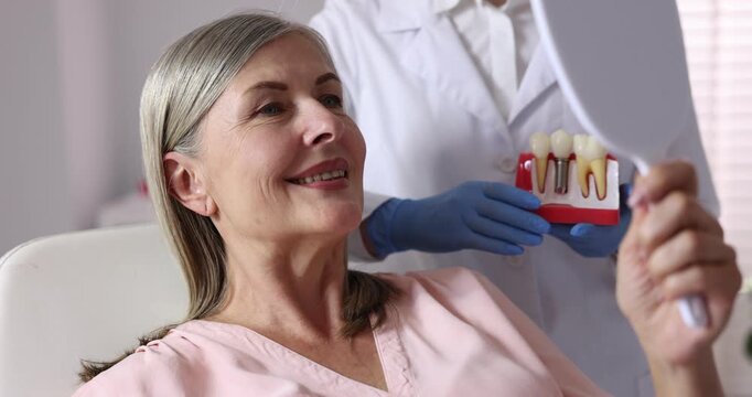 Woman looking at her teeth in mirror and doctor with dental implant model in clinic, closeup