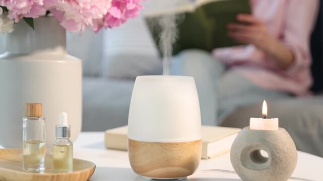 Aromatherapy. Woman reading book at home, selective focus. Modern aroma lamp, burning candles and bottles of essential oils on light table indoors. Camera tilting up