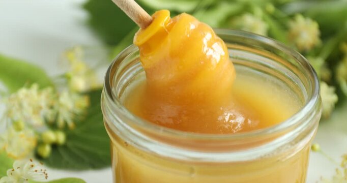 Dripping sweet honey into glass jar and linden flowers at table, closeup