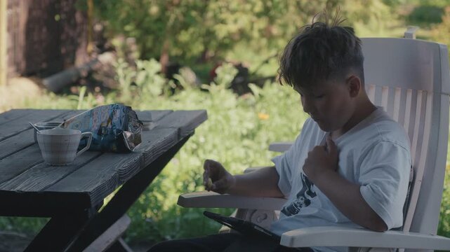 A casual lifestyle scene of a child snacking on crunchy crisps while engrossed in a digital screen outdoors. Great for family tech usage, summer vacation, and childhood lifestyle content.