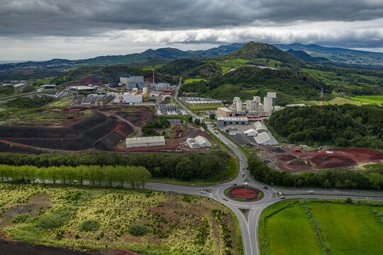 Aerial view of an industrial area with factory buildings, a roundabout, and surrounding green hills under a cloudy sky in Pico da Pedra, Azores, Portugal.