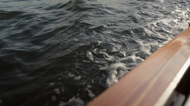 A cinematic close up shot of dark blue water ripples and white foam wake alongside a polished wooden boat hull. Ideal for travel, luxury sailing, and maritime lifestyle content.