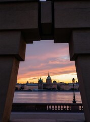 Fototapeta premium Stone monument frames historic cityscape across river at sunset. Pastel sky with clouds. Ornate buildings and classic lamppost visible. Calm water reflects twilight glow.
