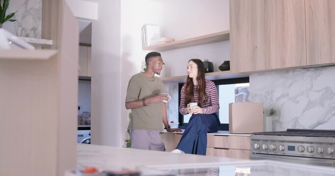 Couple chatting, sipping mugs, clinking cups and laughing amid unpacking boxes in modern kitchen