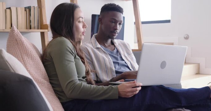 Diverse couple sitting on couch positioning silver laptop, typing and pointing while reviewing work