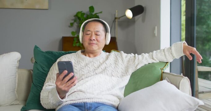 Senior Chinese man in cream sweater and headphones holding phone, scrolling to play audio on sofa