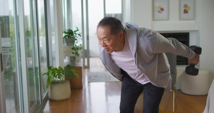 Senior Chinese man holding hex dumbbells, doing bent-over extensions in living room for strength