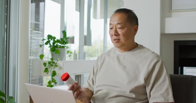 Senior man in tan shirt lifting redcap bottle, studying label and comparing laptop info at home