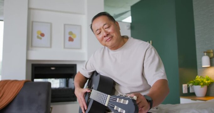 Senior Chinese man tuning black acoustic guitar on bed, hearing off note and adjusting pegs