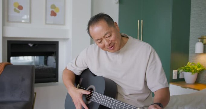 Senior Asian man hearing off note, sitting at home, plucking black guitar, tuning pegs for pitch