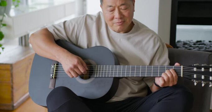 Senior Asian male placing acoustic guitar on lap on couch, plucking chords to relax