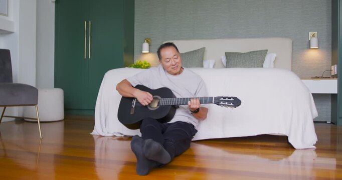 After resting guitar, Senior Chinese man sitting on floor at foot of bed, practicing chords