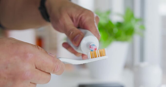 Senior Asian man holding toothbrush squeezing tube at sink, applying tricolor paste for brushing