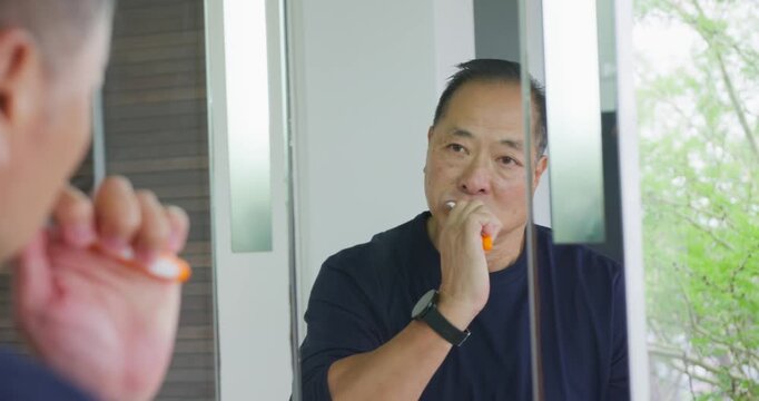 Senior Asian man brushing teeth at mirror for morning routine with orange toothbrush wearing watch
