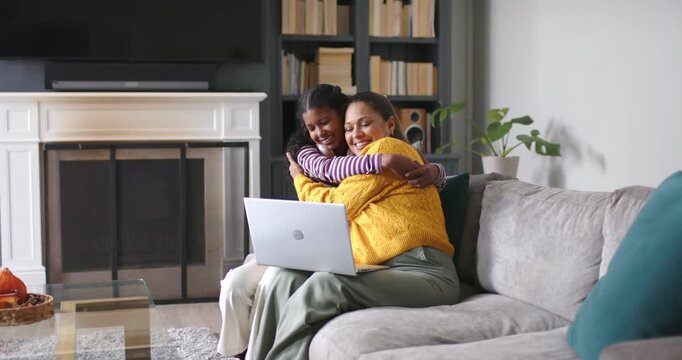 African American mother and teen daughter hugging, daughter leaning to see laptop on sofa, laughing