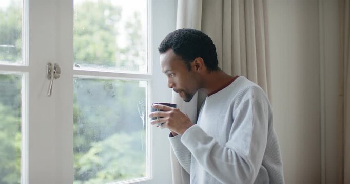African American man in sweater lifting mug, sipping at window, peering out in thought, copy space