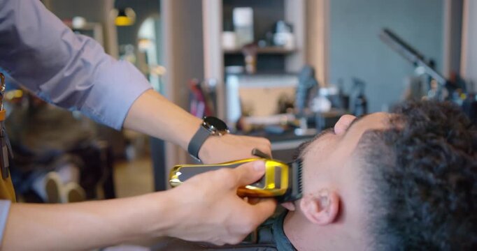 African American barber trimming beard with clipper as male client reclining in shop for neat edge
