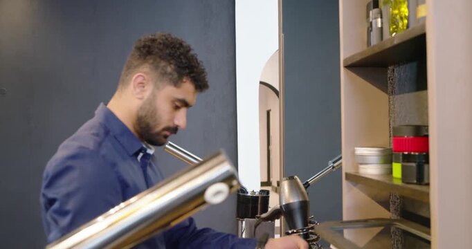 Male stylist standing at salon counter, reaching and adjusting dryer, clippers, prepping for client