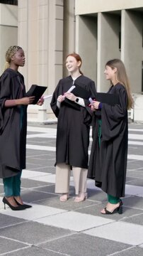 Vertical video: Lifting cap, left grad offering, peers celebrating with diplomas in gowns on plaza