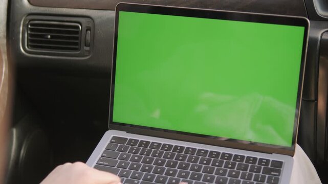 Laptop with green screen inside car. Hand types on keyboard. Person focused on work. Useful for remote work or driving scenes. Green screen for adding content later