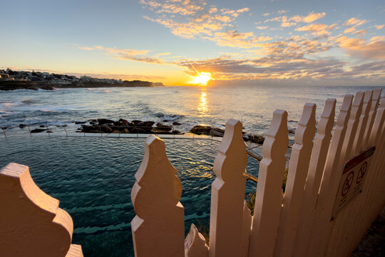 Sunrise over Bronte Ocean Pool viewed through a white picket fence with golden light reflecting across the Pacific Ocean, Bronte Beach, Sydney, Australia.