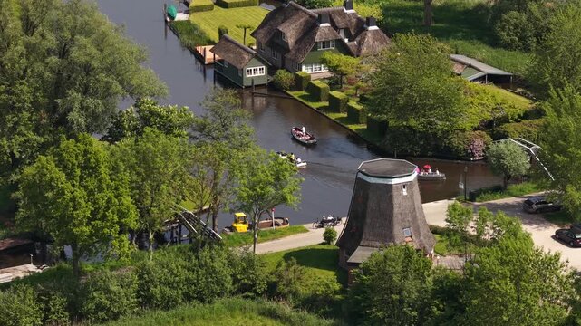 High angle view of tourists enjoying a boat trip along the picturesque canals of giethoorn, passing by traditional thatched roof houses and a historic windmill on a sunny summer day
