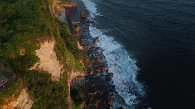 High angle drone shot of steep green cliffs meeting deep blue sea surf in Bali at dusk. Breathtaking tropical island coastline scenery.