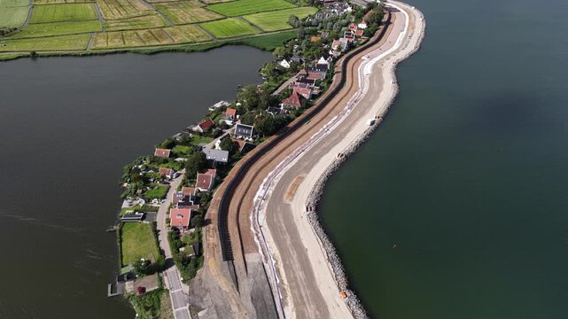 Aerial perspective of ongoing dike reinforcement and coastal protection construction, a massive civil engineering project safeguarding the shoreline village from rising sea levels and flooding