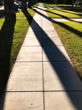 A long sidewalk with diagonal shadows of trees and grass on both sides along a street