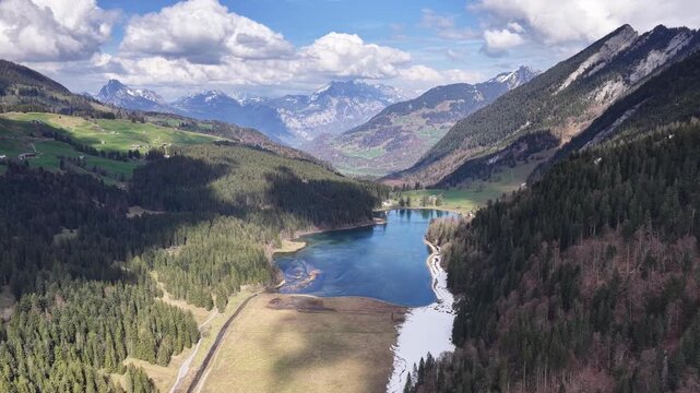 Drone flight above a tranquil alpine lake bordered by forests and mountain slopes in the Swiss Alps. Stunning aerial perspective of pristine nature and crystal-clear water.