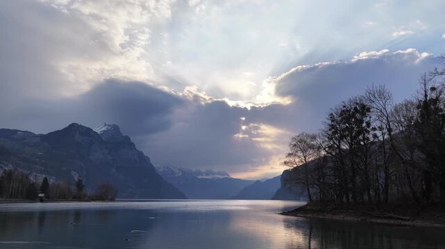 Cinematic drone pullback over Walensee lake in Switzerland, dramatic sun rays through clouds, alpine mountains, calm water reflections and peaceful evening atmosphere.