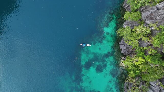 Spectacular top-down aerial drone clip of a lone adventurer paddling a kayak through the transparent turquoise waters of a secluded tropical lagoon hidden by towering limestone cliffs in Palawan.