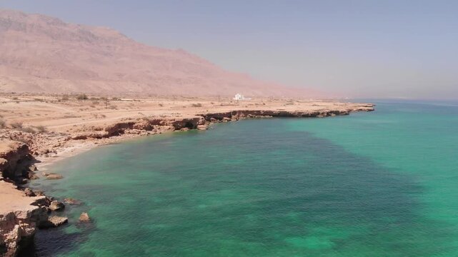 High-angle aerial drone clip of a winding coastal road alongside the turquoise ocean waves and rugged desert cliffs of Oman. Showcases the dramatic contrast of arid landscape and sea.