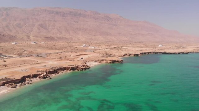 High-angle aerial drone clip of a winding coastal road alongside the turquoise ocean waves and rugged desert cliffs of Oman. Showcases the dramatic contrast of arid landscape and sea.