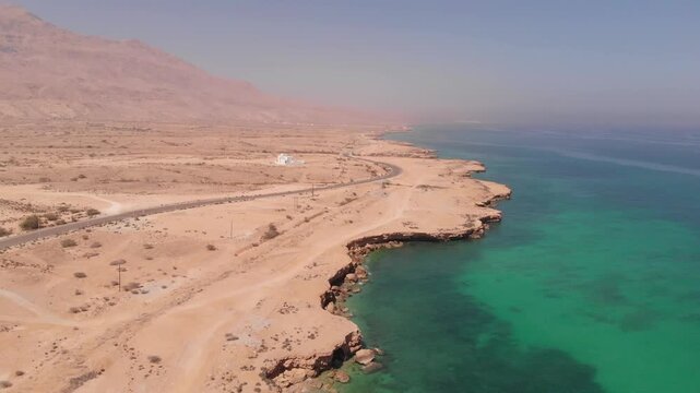 High-angle aerial drone clip of a winding coastal road alongside the turquoise ocean waves and rugged desert cliffs of Oman. Showcases the dramatic contrast of arid landscape and sea.