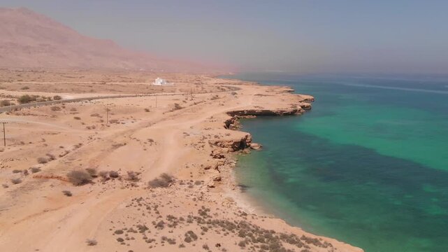 High-angle aerial drone clip of a winding coastal road alongside the turquoise ocean waves and rugged desert cliffs of Oman. Showcases the dramatic contrast of arid landscape and sea.