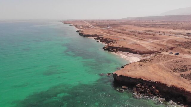 High-angle aerial drone clip of a winding coastal road alongside the turquoise ocean waves and rugged desert cliffs of Oman. Showcases the dramatic contrast of arid landscape and sea.