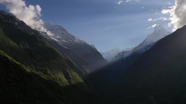 Breathtaking view of the iconic Machapuchare peak and snow-capped Himalayan mountains through a deep, forested valley in Nepal. Features dramatic sunbeams slicing across dark mountain ridges.