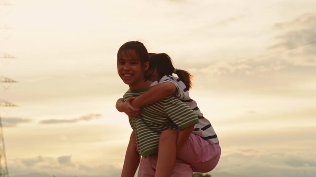 Happy Asian siblings playing together, older sister giving piggyback ride to younger sister at sunset.
