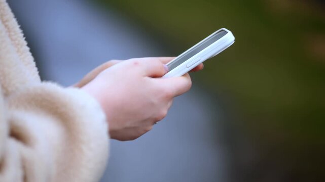 Close-up of hands holding smartphone outdoors. Person wears cozy jacket, focused on device. Blurred background suggests casual, everyday setting. Suitable for themes like digital communication